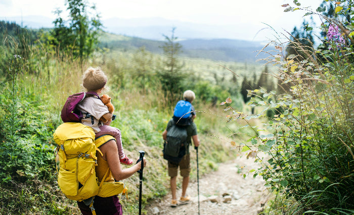 Two people hiking on a trail with backpacks and children, surrounded by nature.