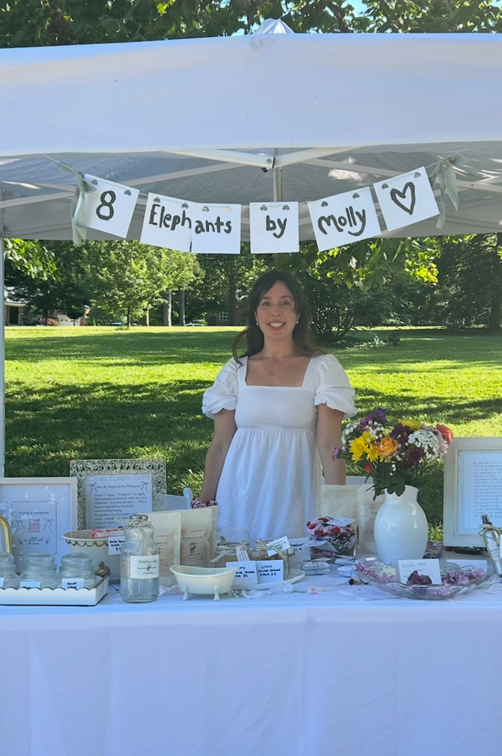 Woman standing behind a table with fresh summer collection, vase of flowers and a display under a white tent in an outdoor setting.