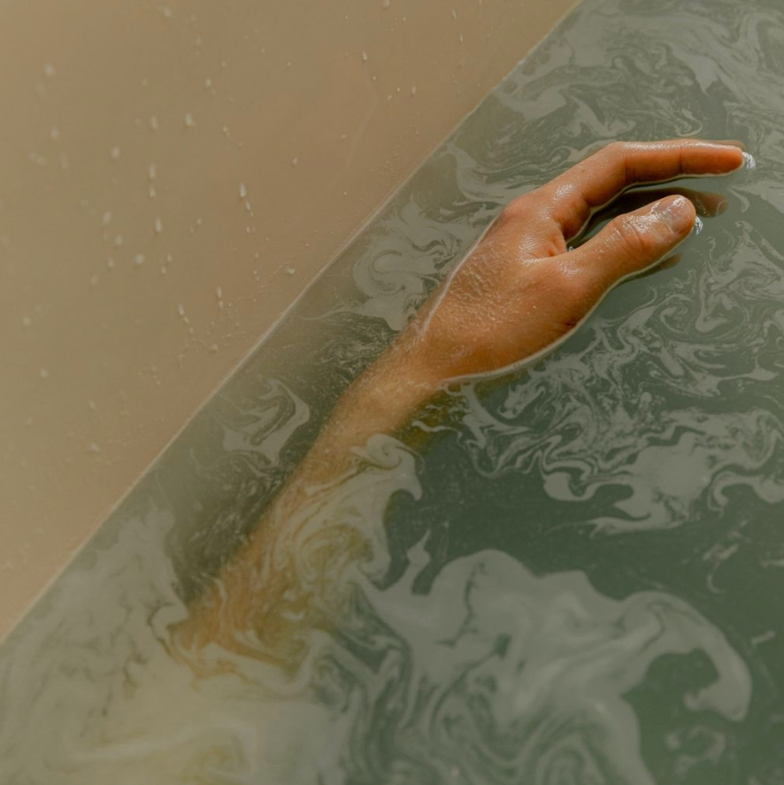 A close-up artistic shot of a person's hand and arm reaching out of murky, foamy water inside a bathtub.
