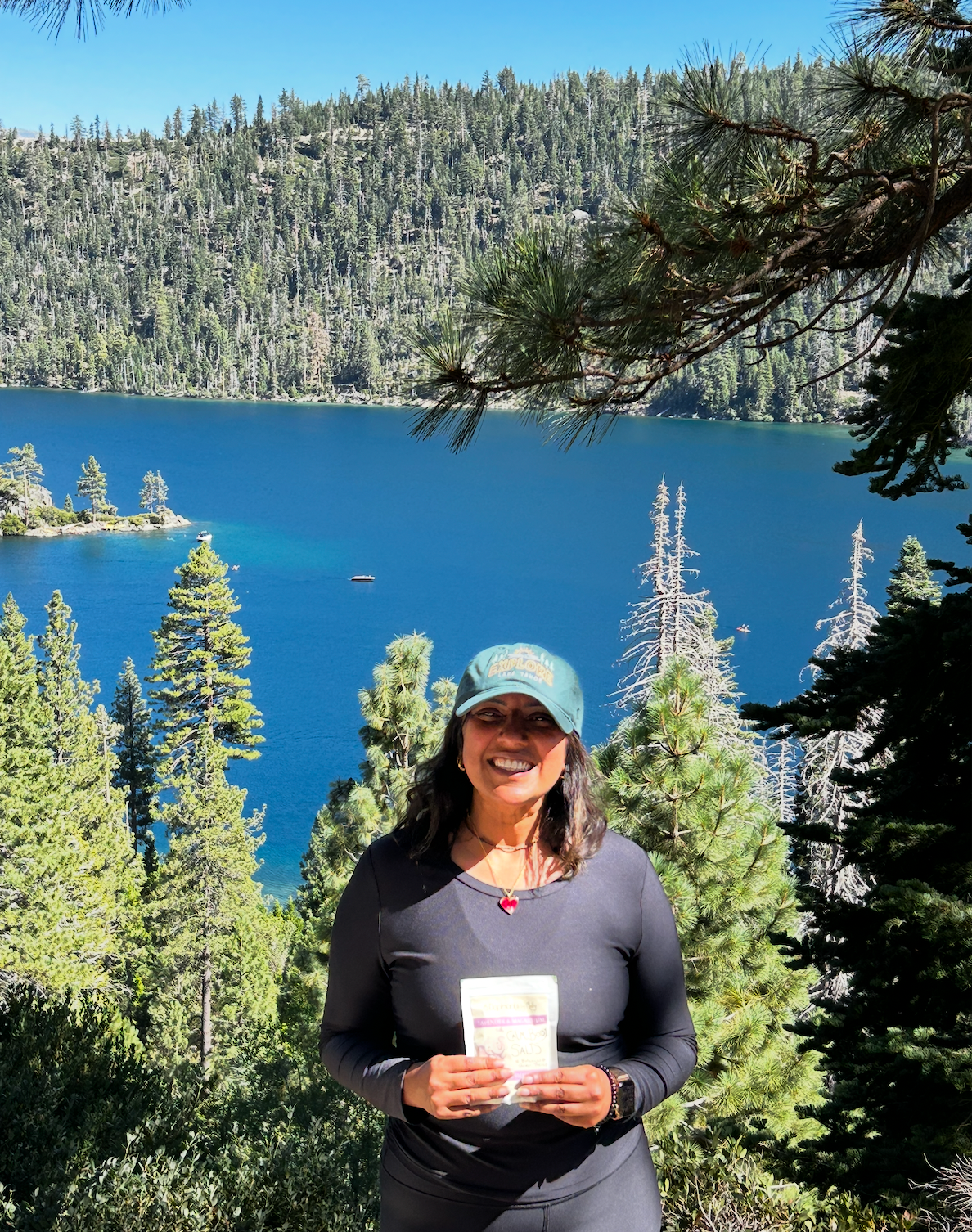 A woman wearing a green cap and black athletic top stands outdoors in front of a blue lake and pine forest, holding a small white product package.