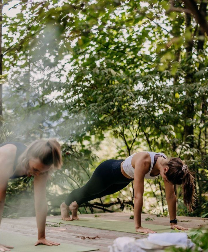 Two women practicing yoga outdoors on a wooden platform with greenery in the background