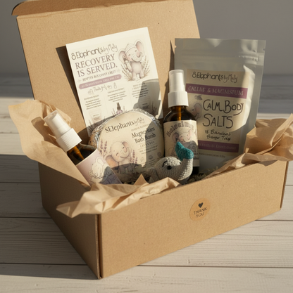 A curated wellness gift box from Elephant by Ella, filled with spa products including a magnesium bath bomb, salts, a spray bottle, and a recovery card, arranged in a brown cardboard box with tissue paper on a light wood surface.